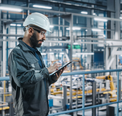 穿着蓝色连身衣和白色安全帽的男子在工厂环境中手持平板电脑