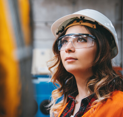 身穿橙色工作背心、戴着安全眼镜和安全帽的年轻女子看着工厂的设备