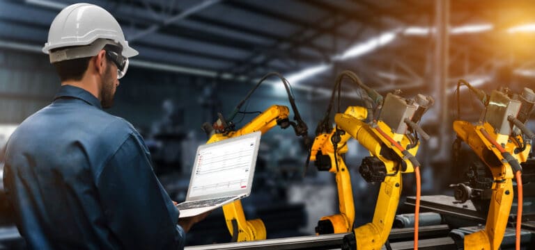 Man standing with laptop next to robotics on the factory floor.