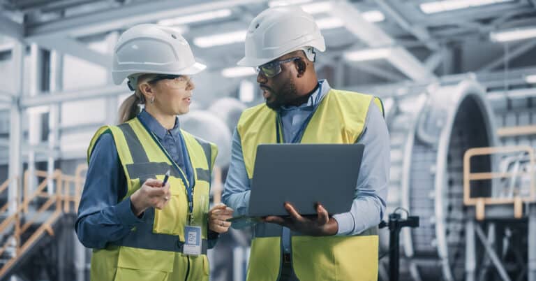 Two Diverse Professional Heavy Industry Engineers Wearing Safety Uniform and Hard Hats Working on Laptop Computer.