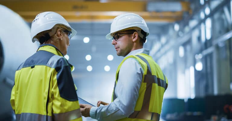 Two heavy industry Engineers stand in pipe manufacturing Factory, using digital tablet Computer, having a discussion.